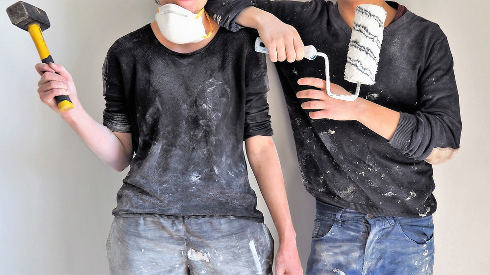 Woman and man in decorating clothing. Man holds a Harris roller while woman is wearing paint splattered clothing; in a dust mask and proudly holding a hammer. 
